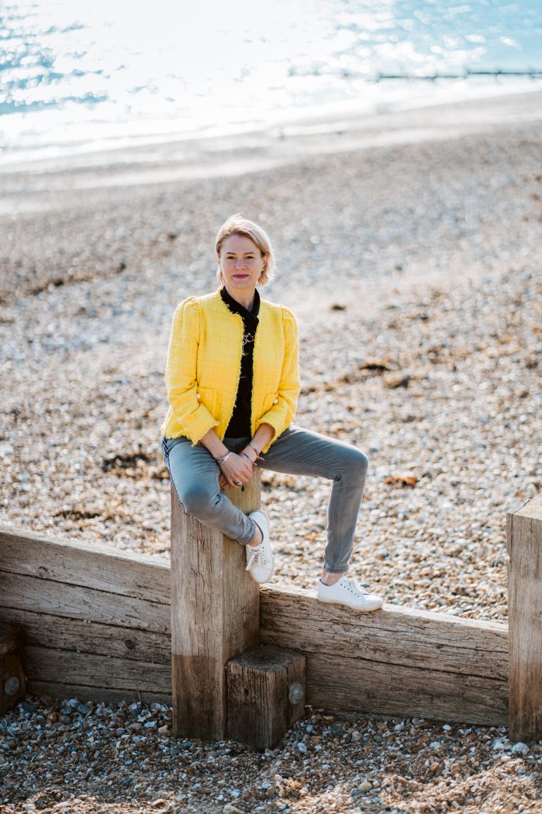 Jane sitting on a groyne on the beach in the sunshine in a yellow jacket